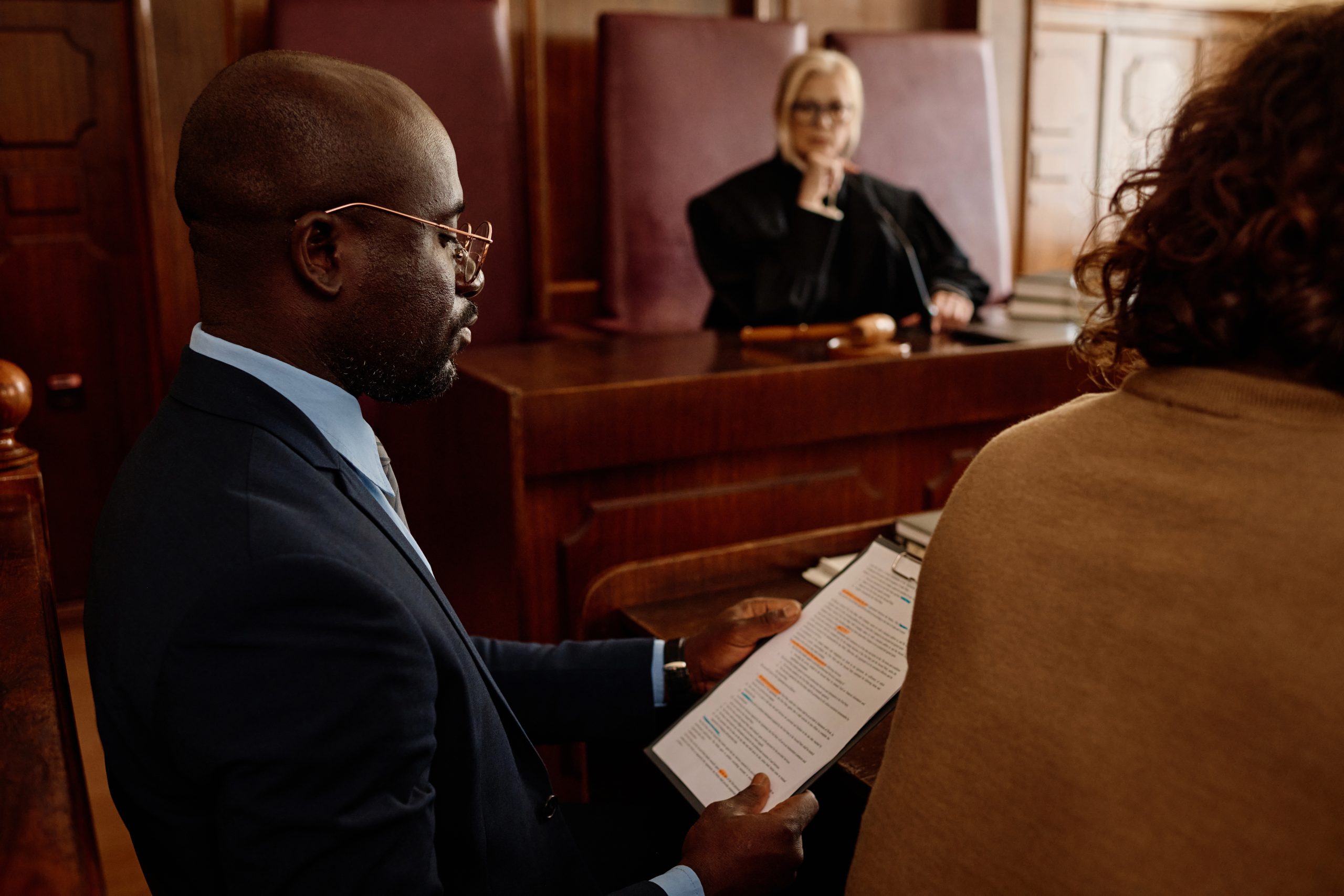 Focus on young confident African American male attorney in formalwear reading juridical document in court while sitting next to suspect